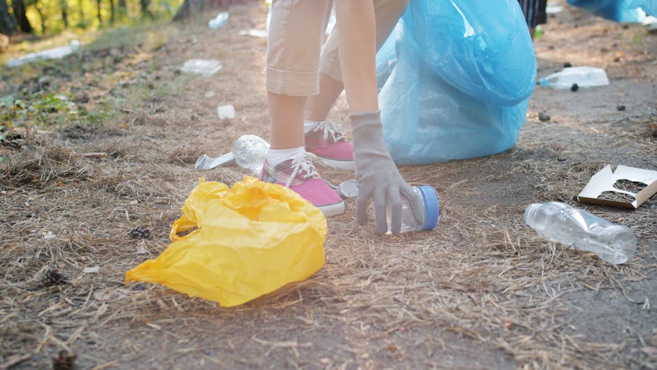 A person wearing white gloves, pink sneakers, and light-colored pants is collecting litter from the ground in an outdoor area during daytime. The ground is covered with dry pine needles and scattered plastic bottles, some crumpled, and a small discarded food container. Nearby, there is a yellow plastic bag filled with trash and a large light blue trash bag. The scene emphasizes environmental cleaning and debris removal, aligning with surface cleaning and hygiene maintenance. The presence of cleaning tools and waste suggests a focus on thorough cleanliness and sanitisation, similar to services offered by Cleaner Haringey for event cleanup in Haringey.
