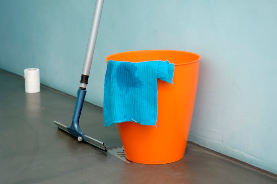 An orange plastic bucket placed on a smooth, grey floor next to a floor drain, with a blue microfiber cloth draped over the edge of the bucket. A mop with a blue handle leans against the wall behind the bucket. In the background, a roll of white paper towels can be seen on the floor near the wall. The scene depicts a setting suitable for surface cleaning and deep cleaning within a commercial or residential space, emphasizing hygiene and sanitation. The wall is painted in a neutral tone, and the area appears clean and well-maintained, reflecting professional cleaning services provided by Cleaner Haringey during event cleanups at Alexandra Palace in Haringey.