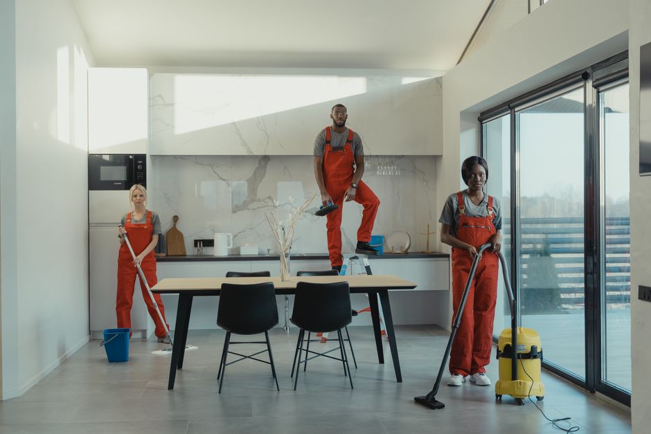 Inside a modern kitchen with white marble walls and large glass sliding doors, three professional cleaners from Cleaner Haringey are engaged in sanitisation and surface cleaning tasks. The female cleaner on the left is standing with a mop and a blue bucket. The male cleaner in the center, elevated on a step ladder, is holding a cleaning cloth, indicating detailed surface cleaning or deep cleaning. The female cleaner on the right is operating a yellow vacuum cleaner, preparing to clean the tiled floor. Natural light fills the room, highlighting the spotless, shiny countertops, ceramic tiles, and wooden dining table with black chairs, reflecting a high standard of hygiene and cleanliness typical of domestic cleaning services. The scene emphasizes thorough surface cleaning and maintenance to ensure a hygienic environment, aligning with the topic of avoiding hidden fees by seeking transparent, quality cleaning services from [COMPANY_NAME].