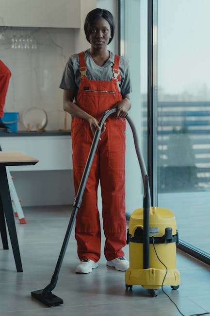 Inside a modern kitchen with white marble walls and large glass sliding doors, three professional cleaners from Cleaner Haringey are engaged in sanitisation and surface cleaning tasks. The female cleaner on the left is standing with a mop and a blue bucket. The male cleaner in the center, elevated on a step ladder, is holding a cleaning cloth, indicating detailed surface cleaning or deep cleaning. The female cleaner on the right is operating a yellow vacuum cleaner, preparing to clean the tiled floor. Natural light fills the room, highlighting the spotless, shiny countertops, ceramic tiles, and wooden dining table with black chairs, reflecting a high standard of hygiene and cleanliness typical of domestic cleaning services. The scene emphasizes thorough surface cleaning and maintenance to ensure a hygienic environment, aligning with the topic of avoiding hidden fees by seeking transparent, quality cleaning services from [COMPANY_NAME].