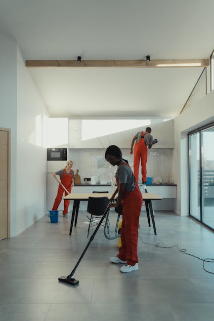 A team of professional cleaners from Cleaner Haringey performing deep cleaning in a modern, spacious kitchen with white walls and large glass sliding doors. The cleaner in the foreground, a woman, is vacuuming the light grey tiled floor with an upright vacuum cleaner. Two other cleaners, wearing red overalls, are at the back: one is scrubbing the kitchen walls near the countertop, while the other is standing on a step ladder, cleaning the upper section of a wall or cupboard. The kitchen features a sleek, minimalist design with stainless steel appliances and a white countertop. Natural light floods the room through the large glass doors, highlighting the cleanliness and shine of the surfaces, emphasizing hygiene and maintenance. This image showcases the thorough approach to surface cleaning, sanitisation, and domestic cleaning services provided by Cleaner Haringey to ensure a hygienic and spotless environment.