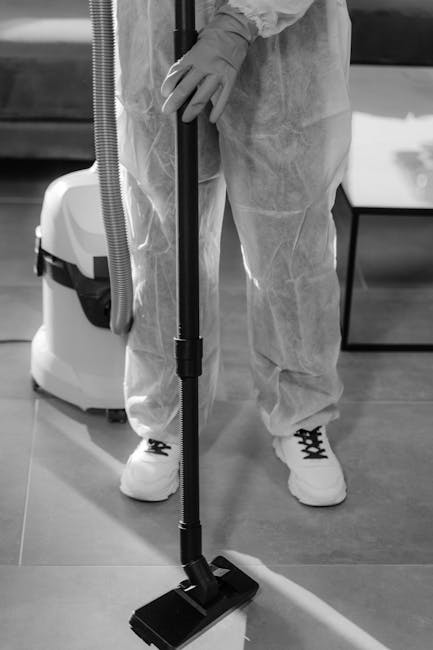 A person wearing protective gloves and a disposable suit is using a black floor vacuum cleaner with a long handle on a smooth, polished wooden floor in a clean, modern room. The vacuum is connected to a white cleaning unit positioned nearby. The room features a dark-colored sofa or chair in the background, and the lighting is bright, highlighting the spotless, dust-free surface. The scene illustrates professional surface cleaning with equipment used by Cleaner Haringey for deep cleaning and sanitisation in residential or commercial settings.
