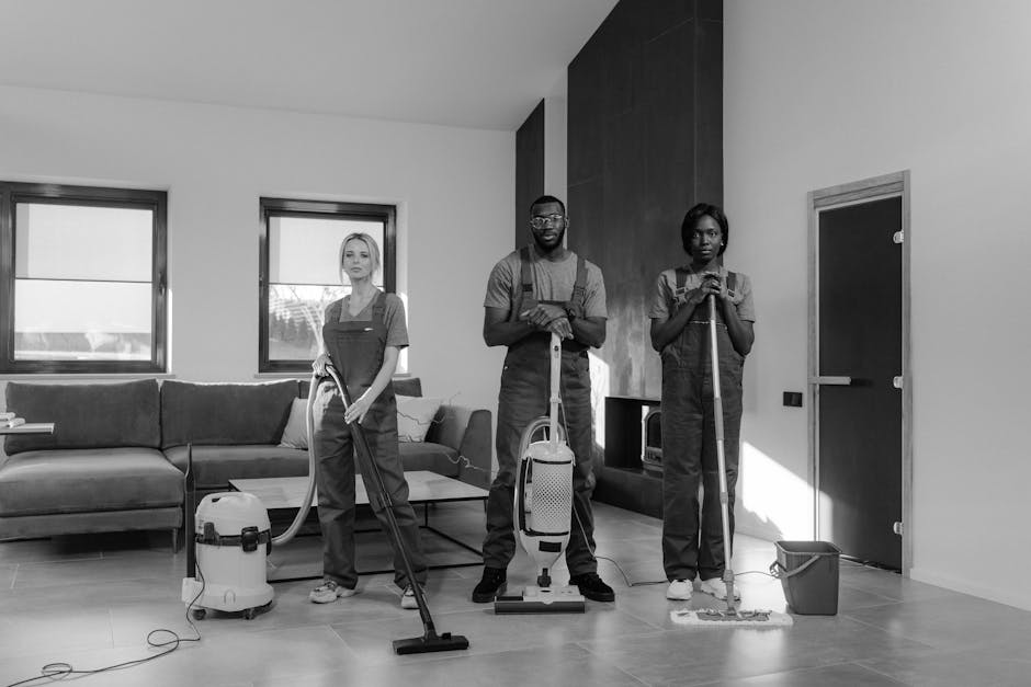 A team of professional cleaners from Cleaner Haringey performing deep cleaning in a modern, spacious kitchen with white walls and large glass sliding doors. The cleaner in the foreground, a woman, is vacuuming the light grey tiled floor with an upright vacuum cleaner. Two other cleaners, wearing red overalls, are at the back: one is scrubbing the kitchen walls near the countertop, while the other is standing on a step ladder, cleaning the upper section of a wall or cupboard. The kitchen features a sleek, minimalist design with stainless steel appliances and a white countertop. Natural light floods the room through the large glass doors, highlighting the cleanliness and shine of the surfaces, emphasizing hygiene and maintenance. This image showcases the thorough approach to surface cleaning, sanitisation, and domestic cleaning services provided by Cleaner Haringey to ensure a hygienic and spotless environment.