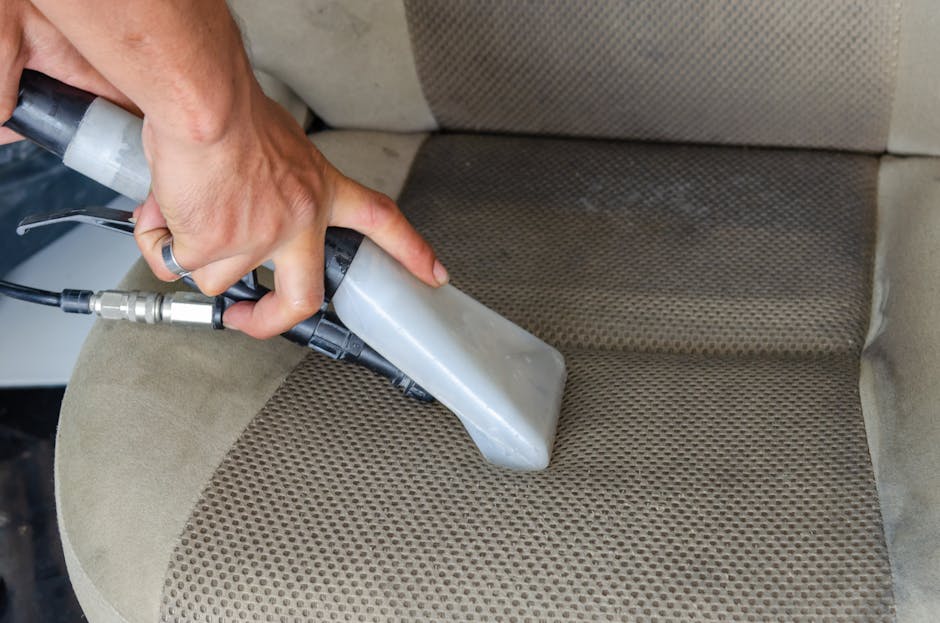 A professional cleaner's hand holding a handheld upholstery cleaning device, applying deep cleaning and sanitisation to a fabric car seat with a beige and brown patterned surface. The device has a black and grey body and is connected to a hose, with visible water or cleaning solution being applied to the fabric. The setting appears to be the interior of a vehicle or a furniture piece, with the surrounding fabric looking slightly stained but actively cleaned. Good lighting highlights the textured surface of the fabric, emphasizing the cleaning process by Cleaner Haringey in their residential or commercial surface cleaning services.