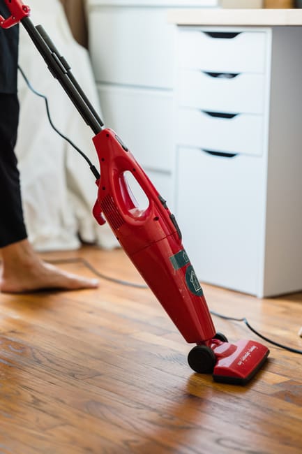 A person using a red handheld vacuum cleaner to clean a wooden floor in a residential room. The scene includes a white storage unit with multiple drawers in the background, and the lighting suggests natural daylight. The vacuum cleaner's nozzle is in contact with the polished wooden surface, which appears clean and free of dust or debris. The individual is barefoot, indicating a domestic setting, and the overall environment reflects a tidy, well-maintained living space. The image showcases surface cleaning and maintenance typical of domestic cleaning services provided by Cleaner Haringey, focusing on deep cleaning and hygiene enhancement.